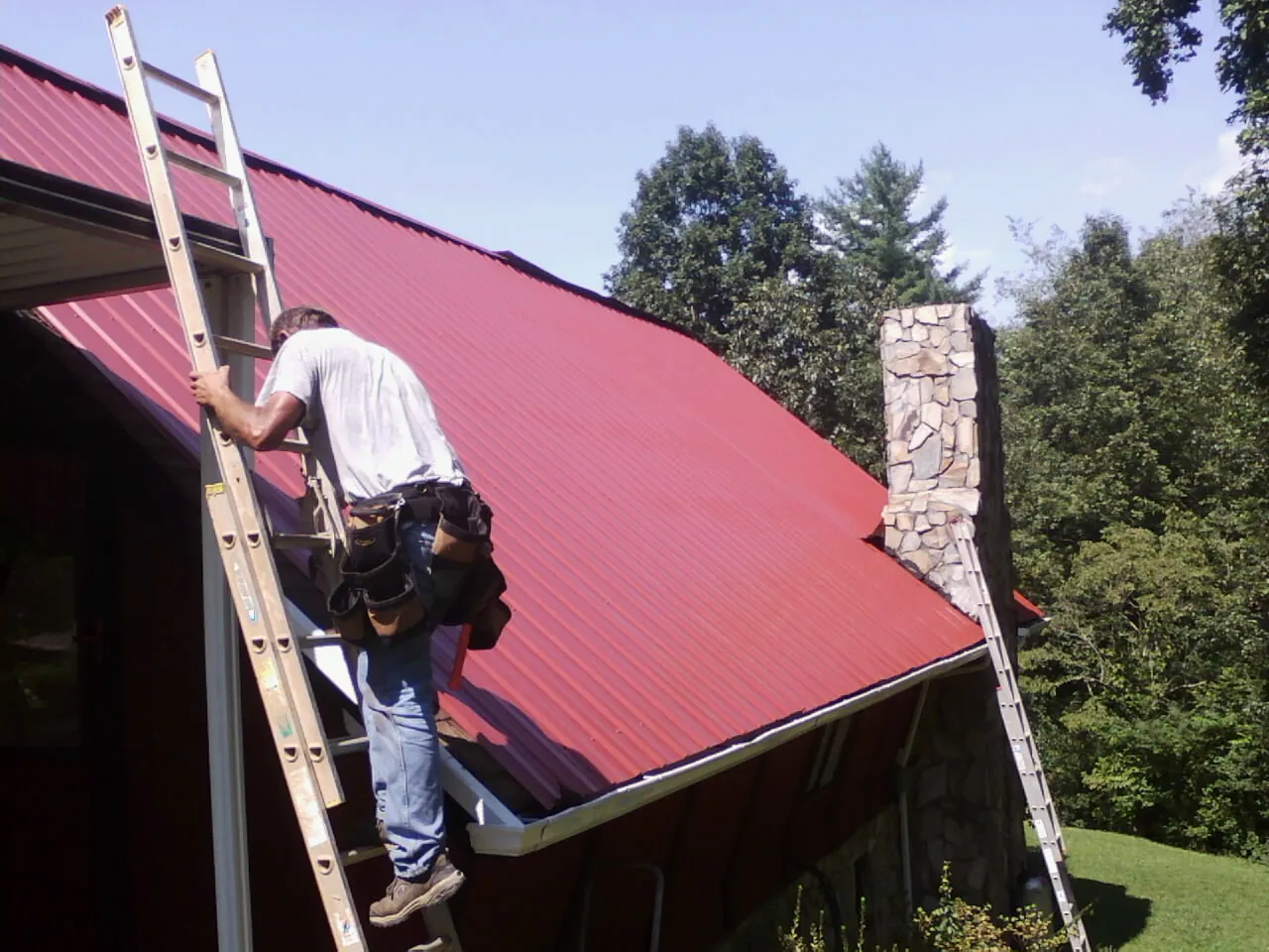 Roofer on a ladder beside a red metal roof and stone chimney — Soffit & Fascia Repair by Roofing Brothers in Bladenboro